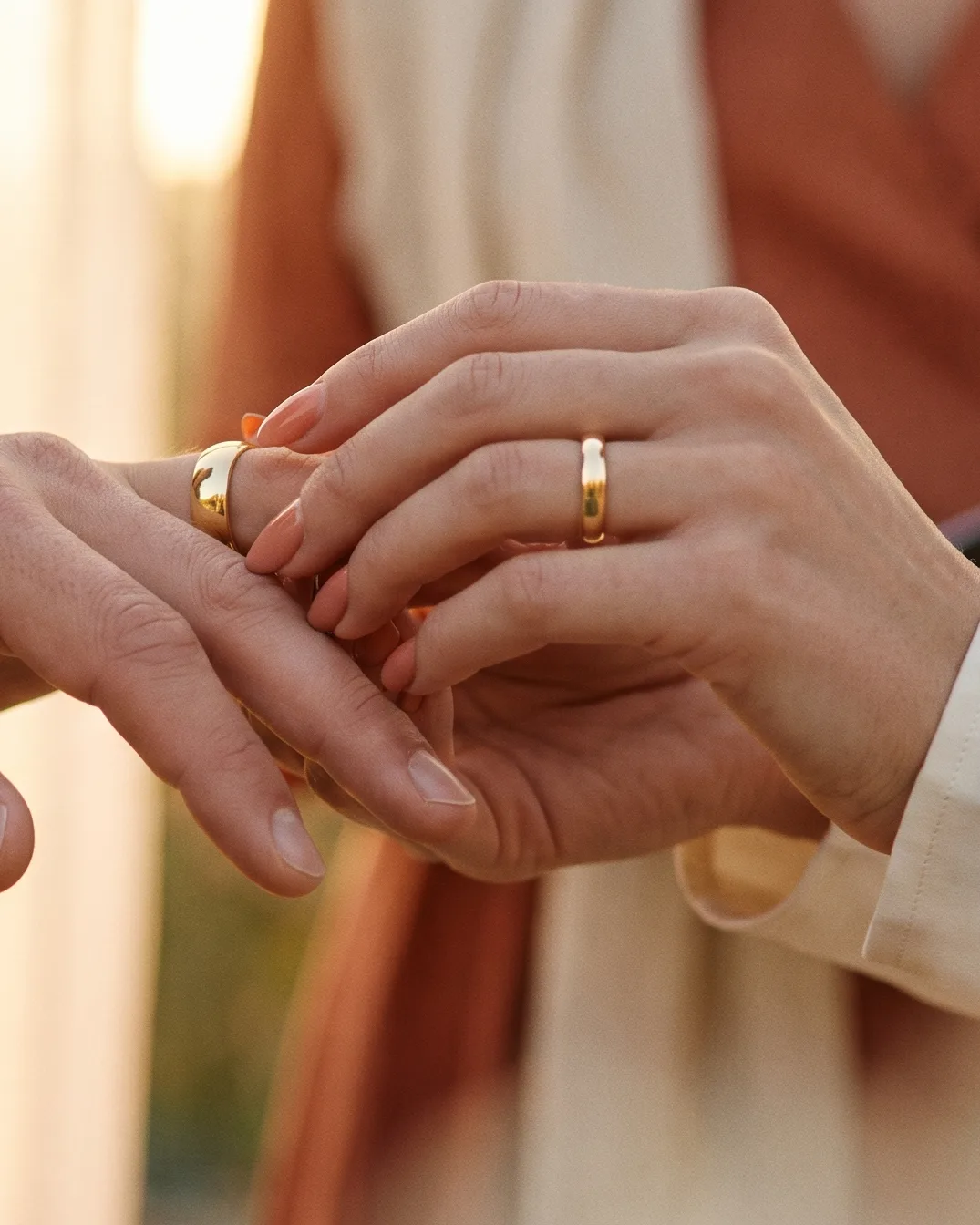 Dos manos entrelazadas al atardecer, cada una con su argolla de matrimonio en oro